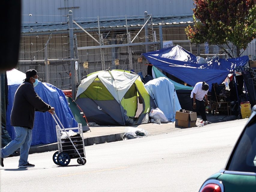 A person pushes a cart in front of numerous tents and makeshift shelters lining a sidewalk, with another individual bending over among the tents. A tree and a fence are in the background.