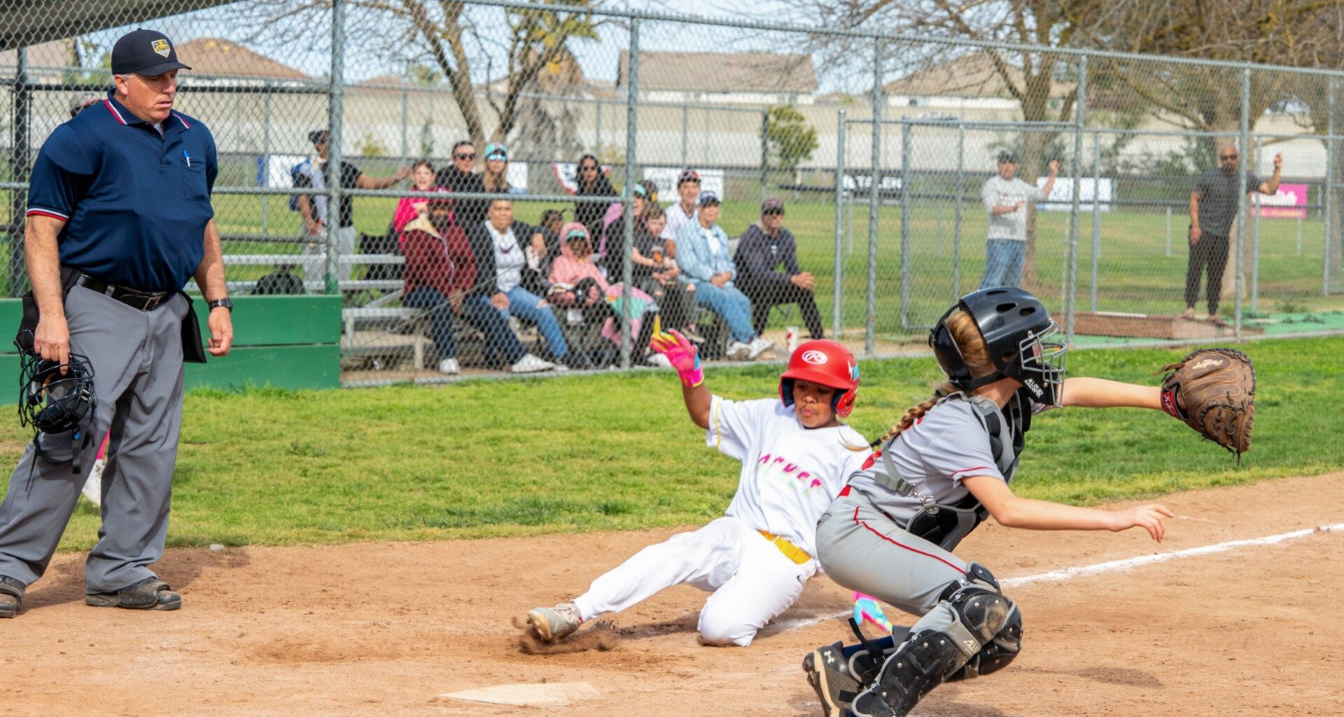 Young girl slides to homebase as umpire watches from side and catcher squats in anticipation of a throw.