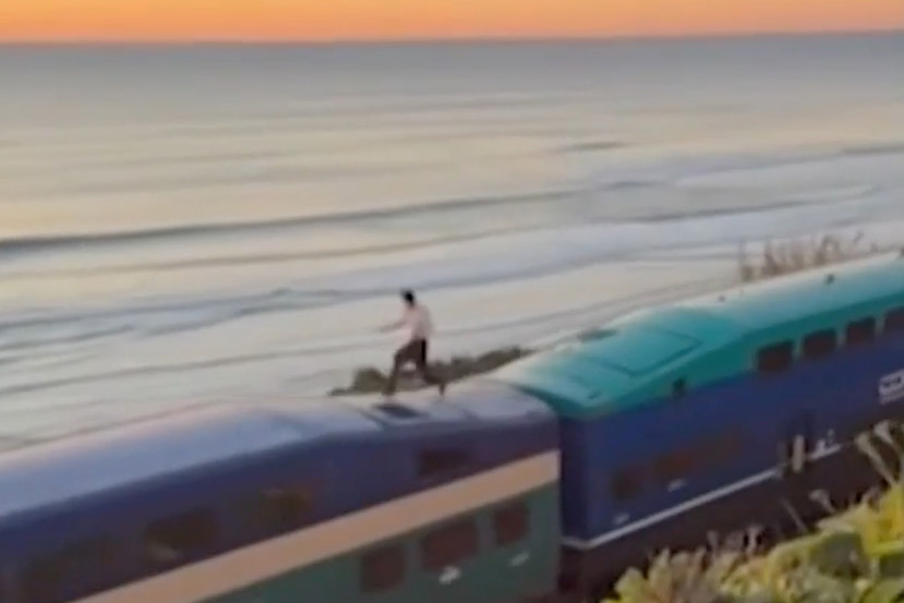 A person appears to be "surfing" atop a moving train next to the ocean at sunset.