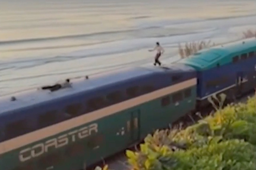 Two people on top of a moving Coaster train near the ocean, one standing and one lying down.