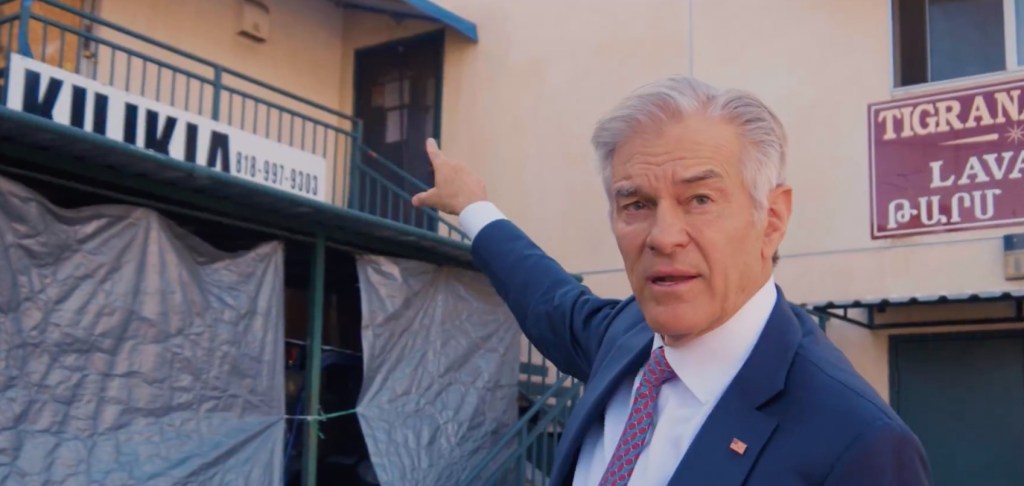Dr. Oz pointing at a building with "KILIKIA" and a phone number on its sign, and another sign for "TIGRANA LAVA."