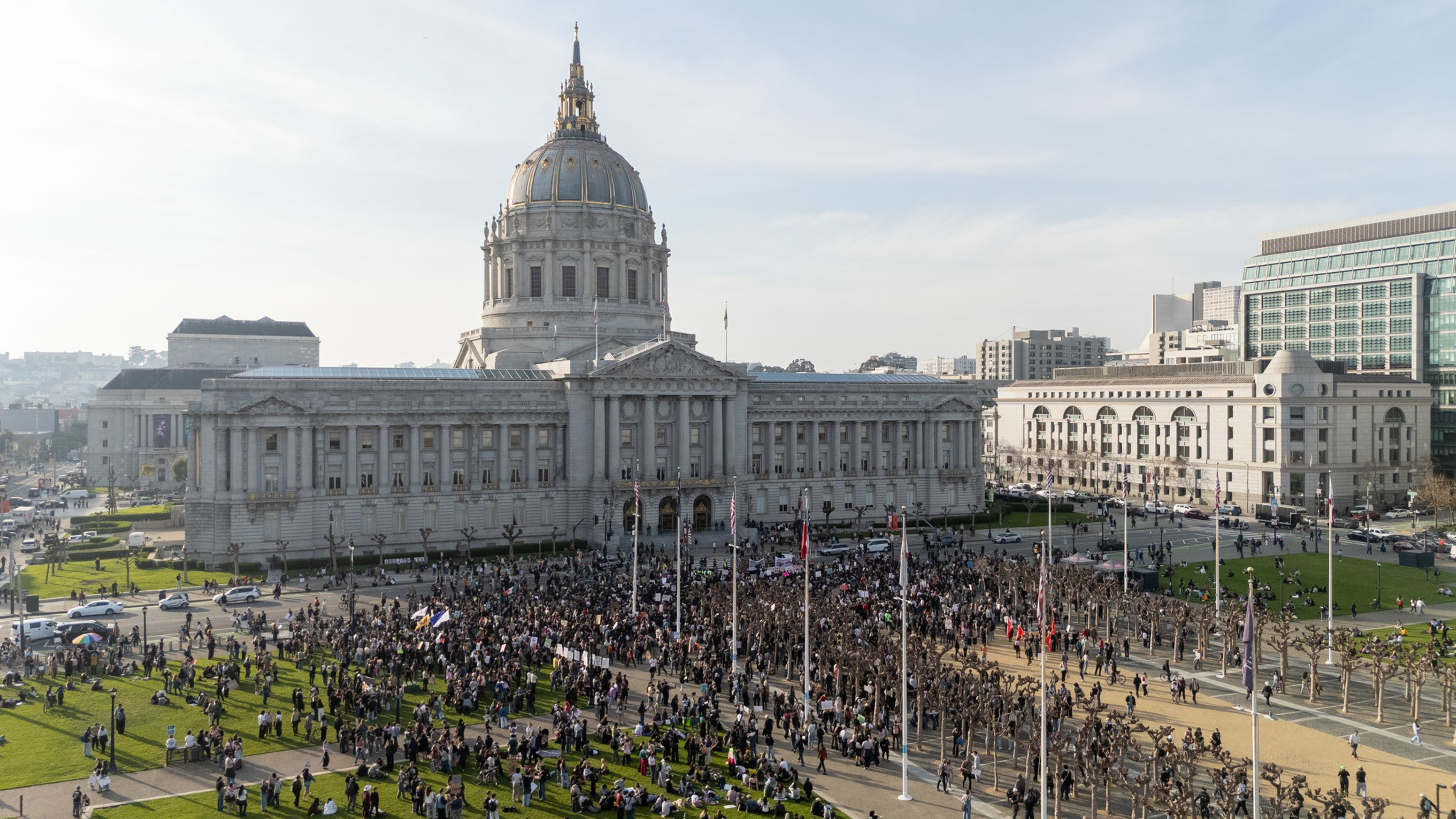 A large crowd gathers on the grass and pavement in front of a grand neoclassical building with a tall domed tower under a clear sky.