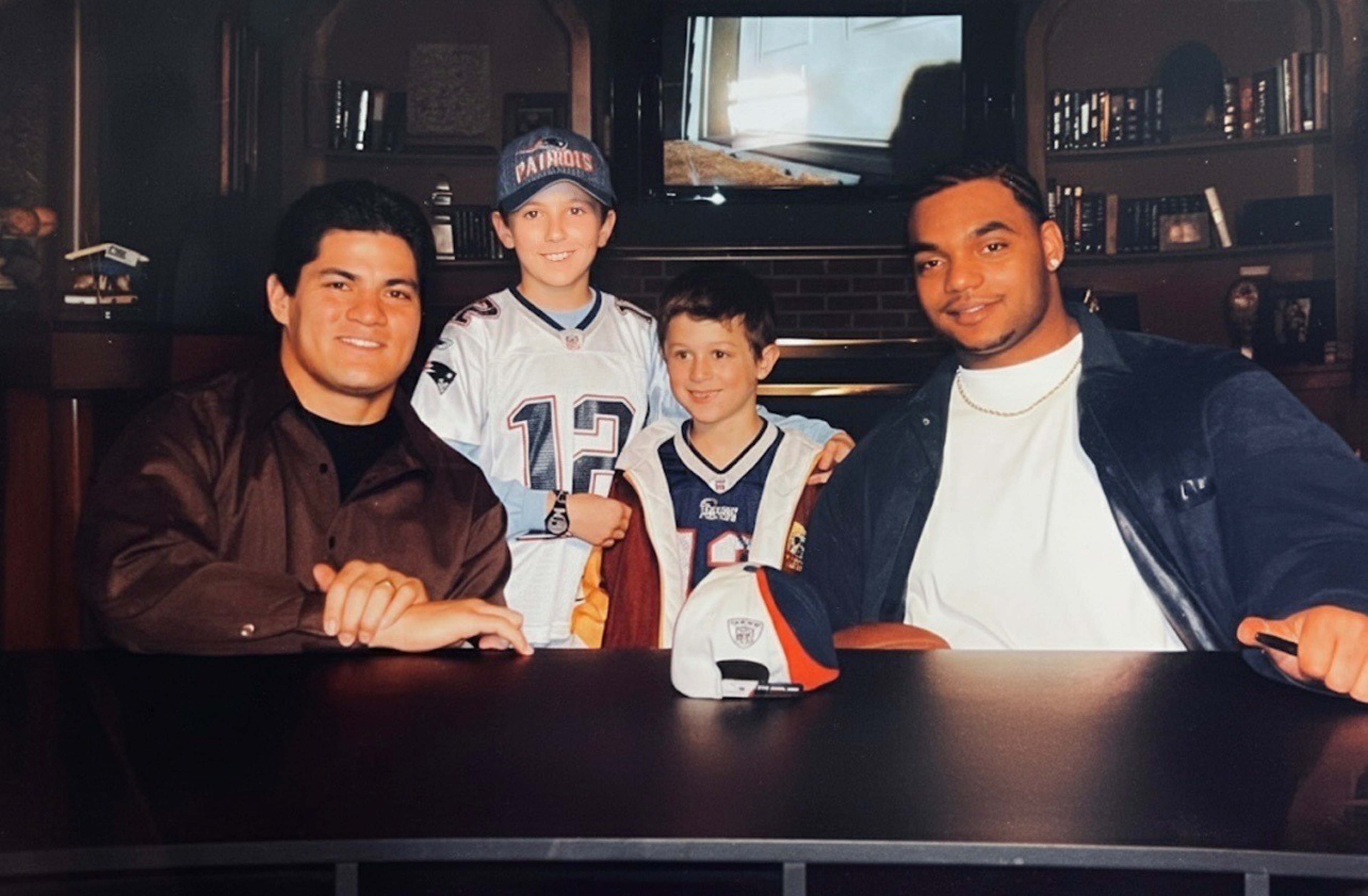 Two young boys wearing New England Patriots jerseys stand behind a table, flanked by two men sitting and smiling, with a sports cap on the table.