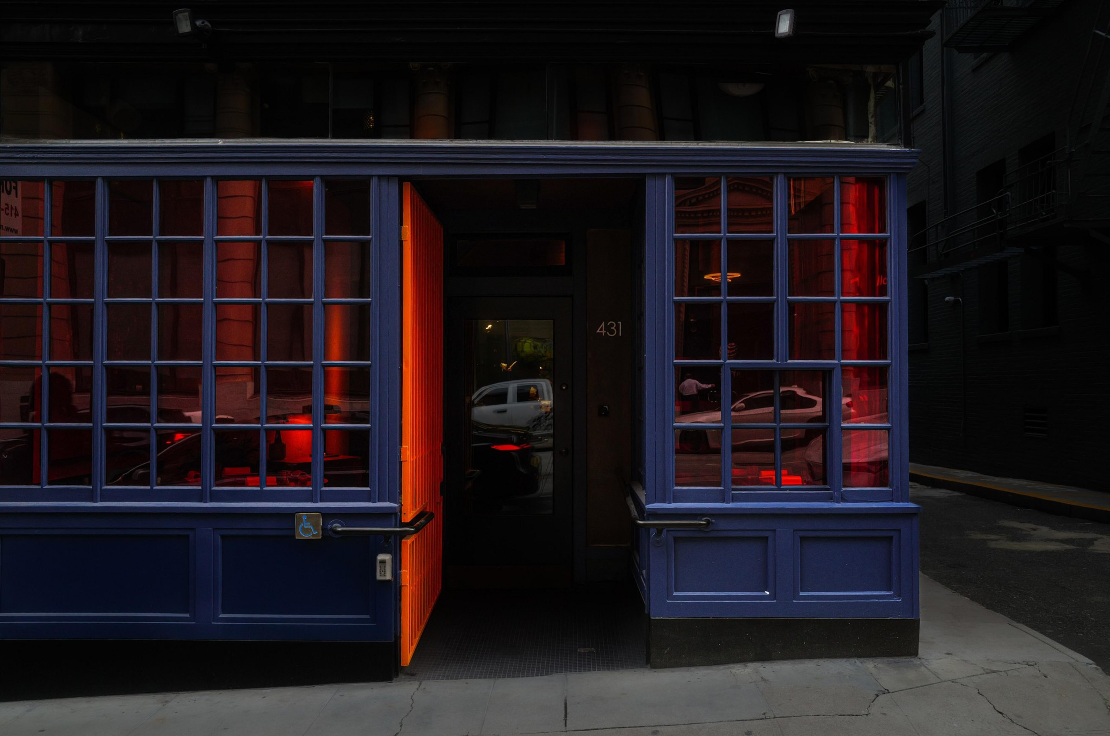 A dark blue storefront with large windows reflects red light; one bright orange door is open, revealing a dim interior with the number 431 beside the door.