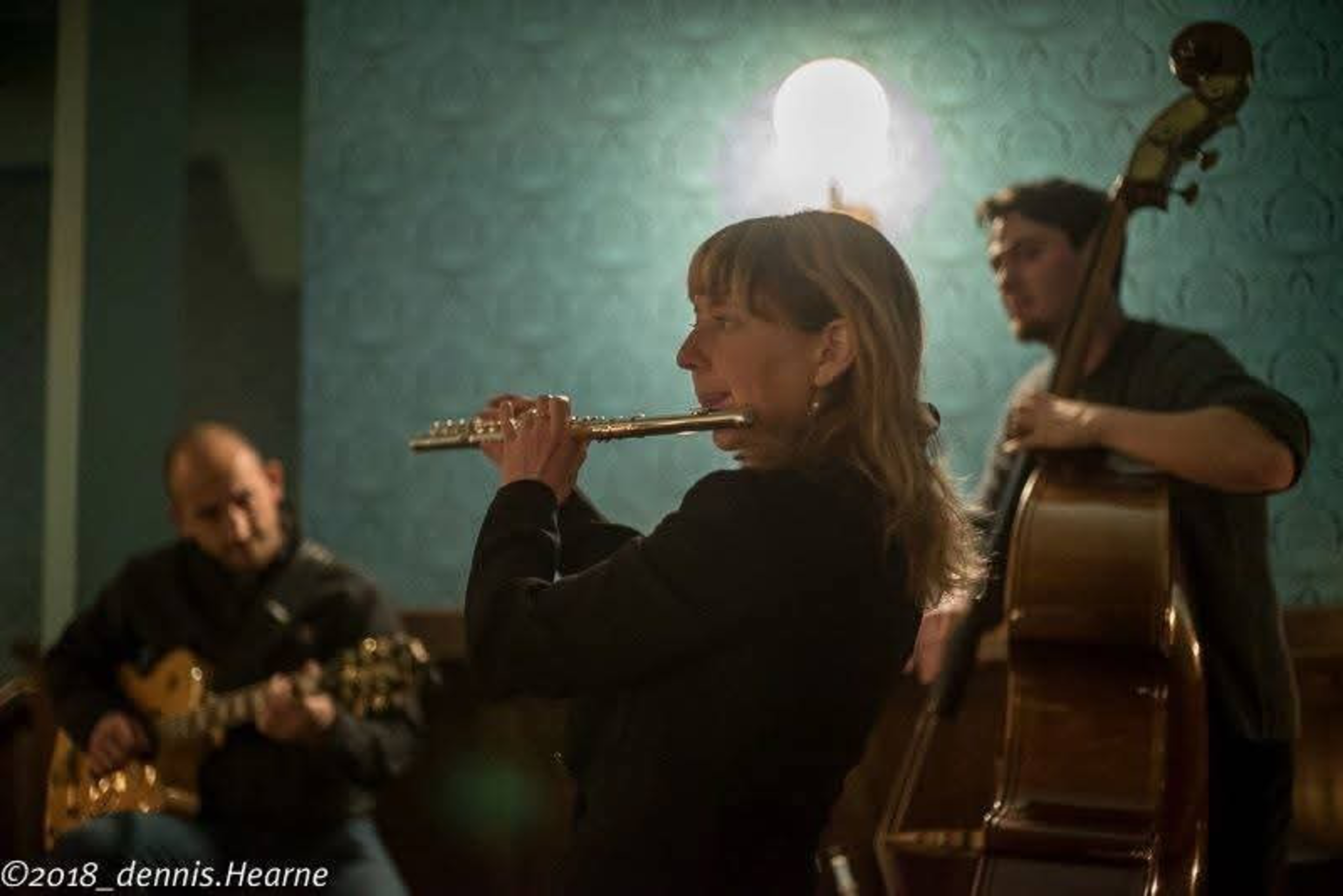 A woman plays the flute while a man plays guitar and another man plays double bass in a dimly lit room.