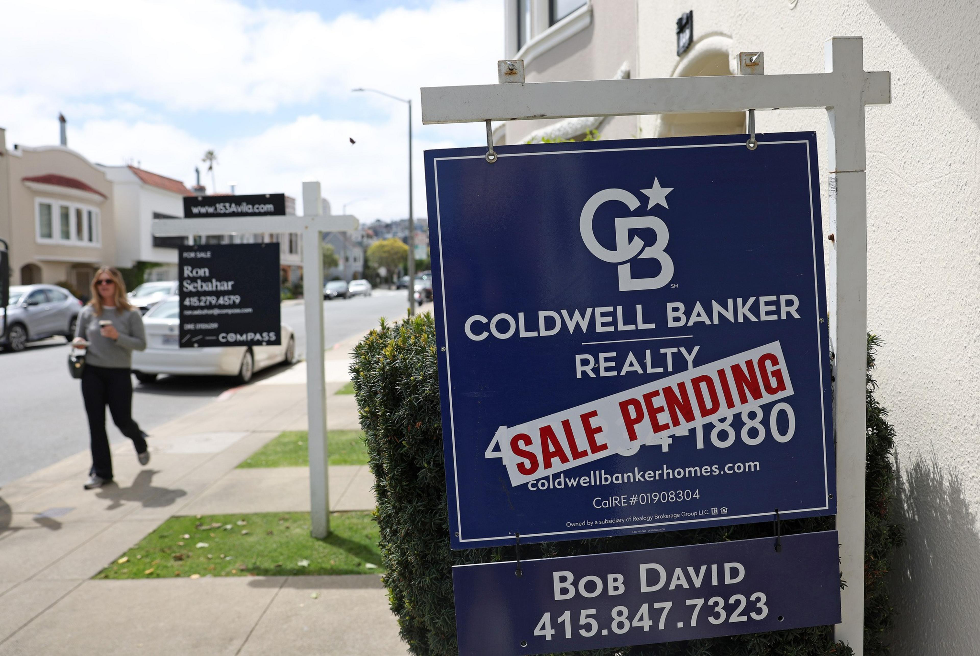 A Coldwell Banker Realty sign with “SALE PENDING” in red covers part of the phone number, visible on a sunny suburban street with a woman walking and another real estate sign in the background.