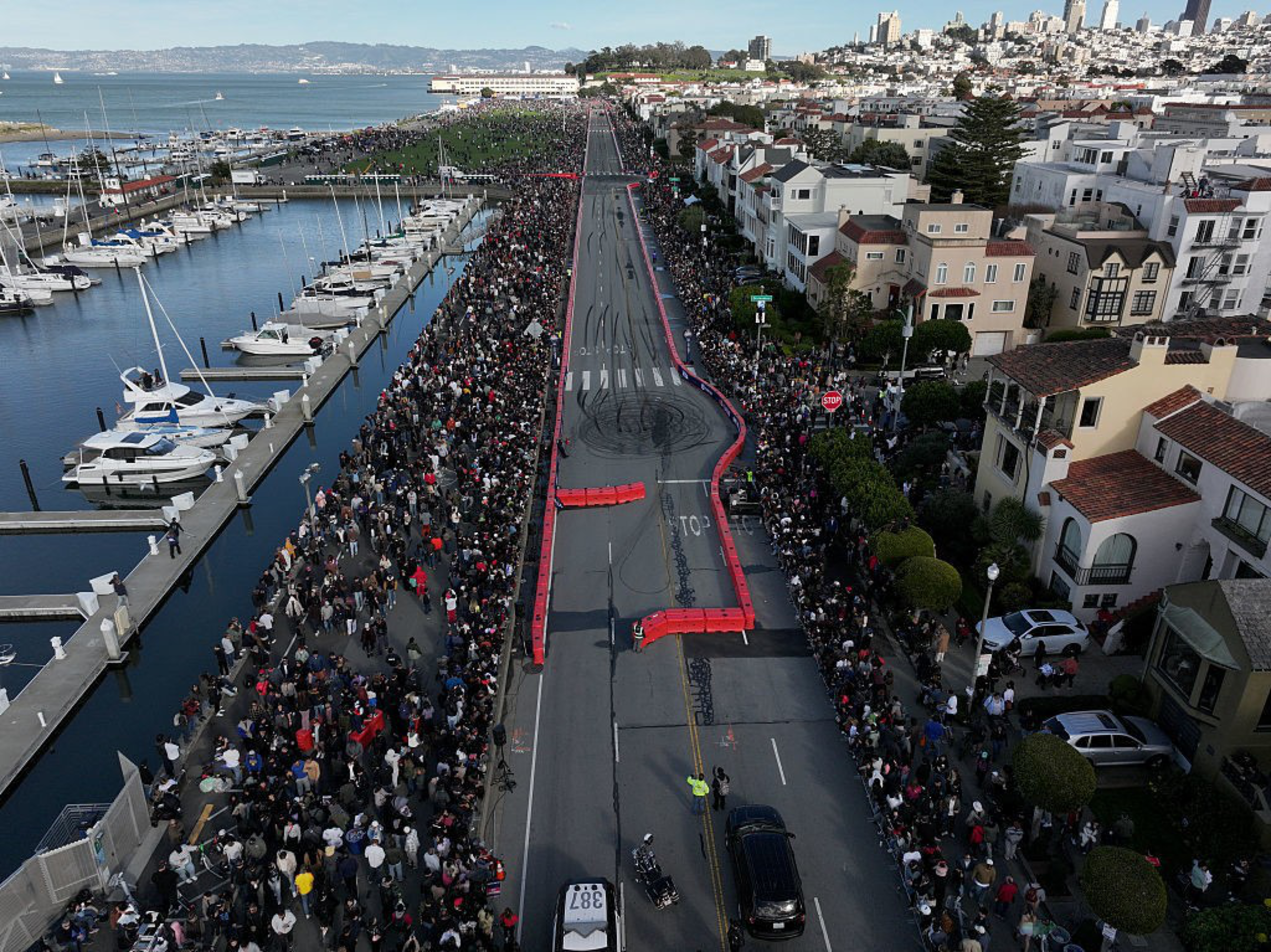 Thousands of people line both sides of a closed coastal street near boats and houses, gathered along barriers with tire marks visible on the road.