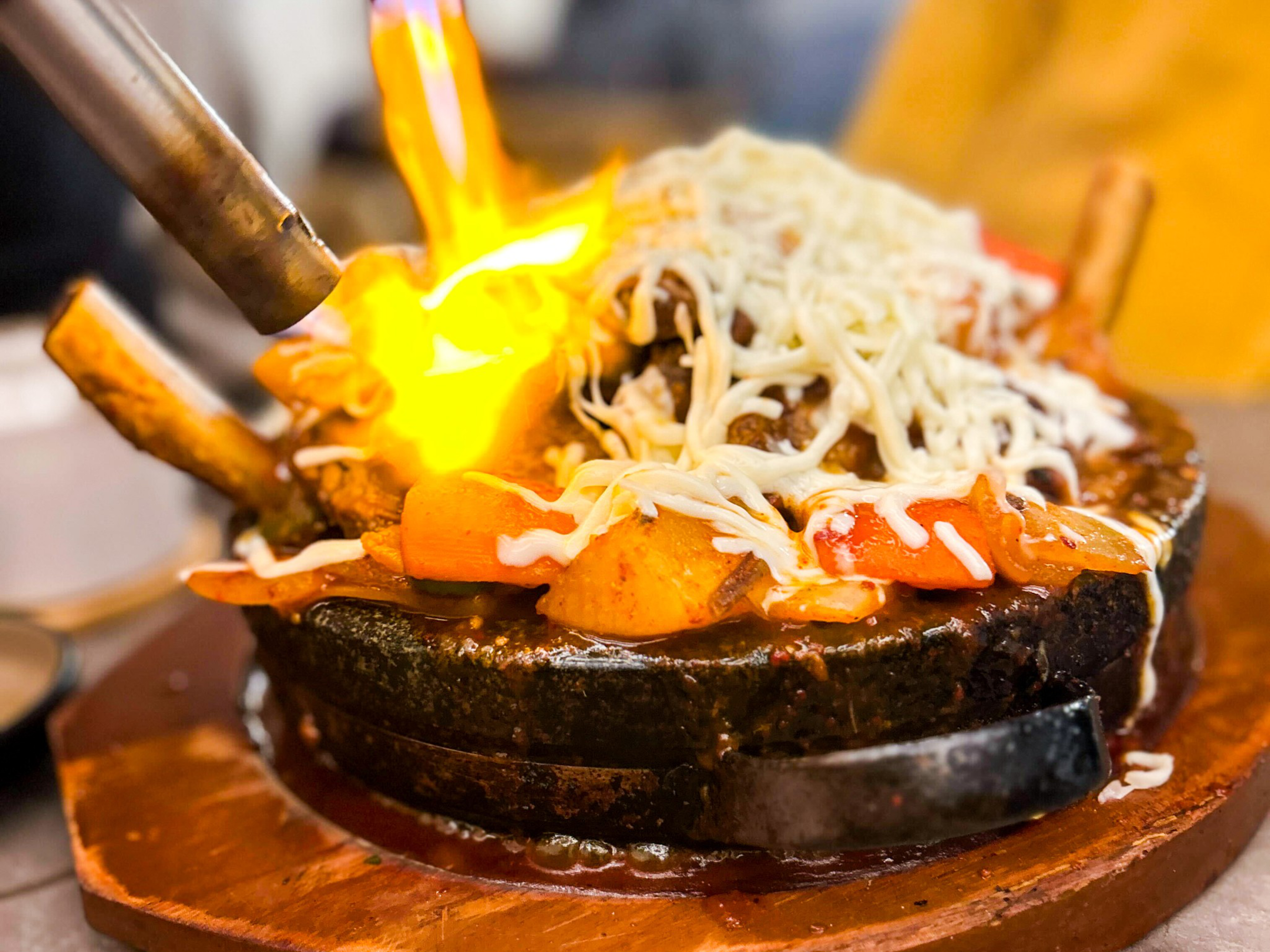 A sizzling dish with meat, vegetables, and melted cheese is being flambéed with a blowtorch on a hot cast iron plate.