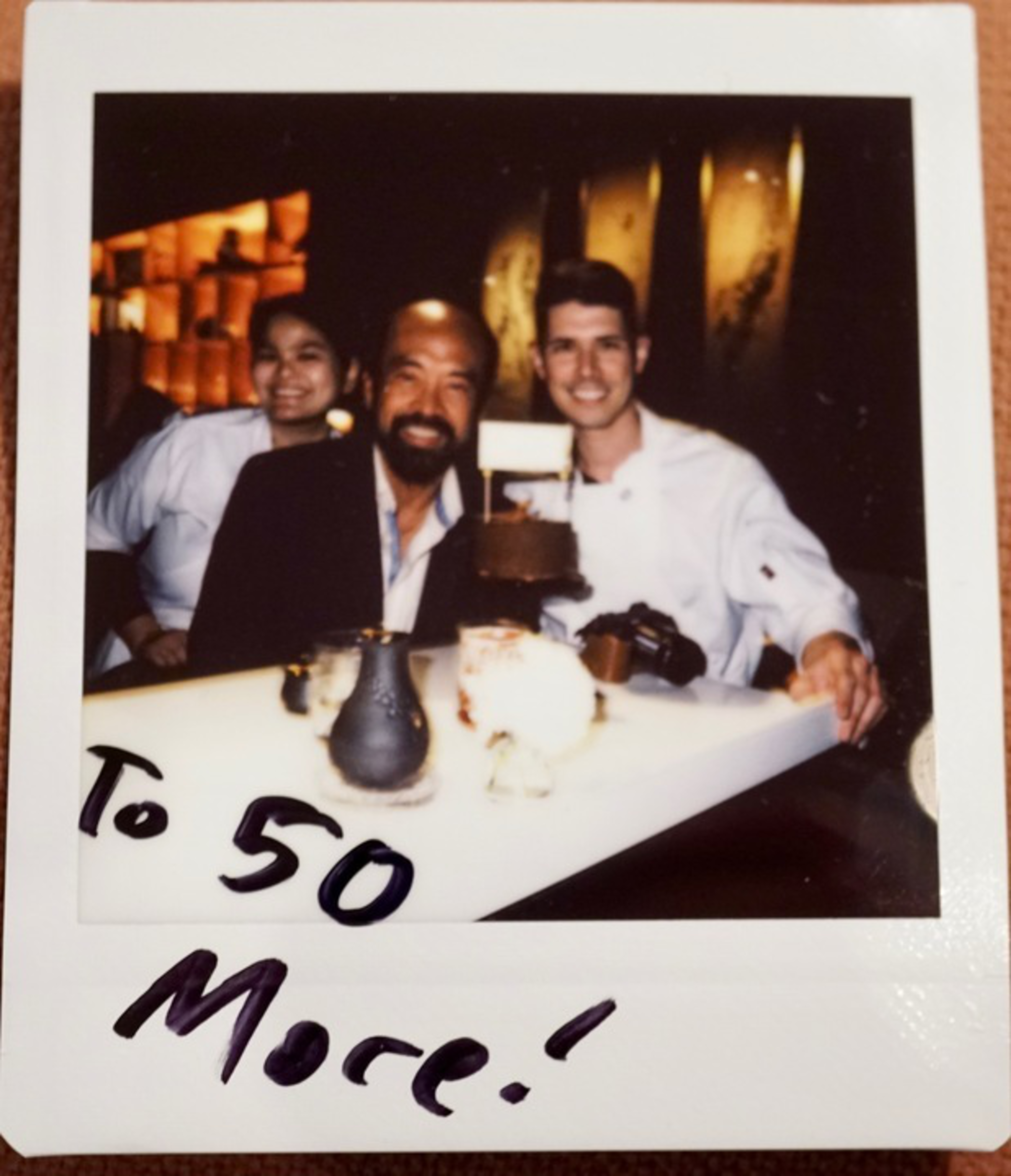 Three people sit at a dimly lit table, smiling, with a small cake on the table; “To 50 More!” is handwritten at the bottom of the photo.