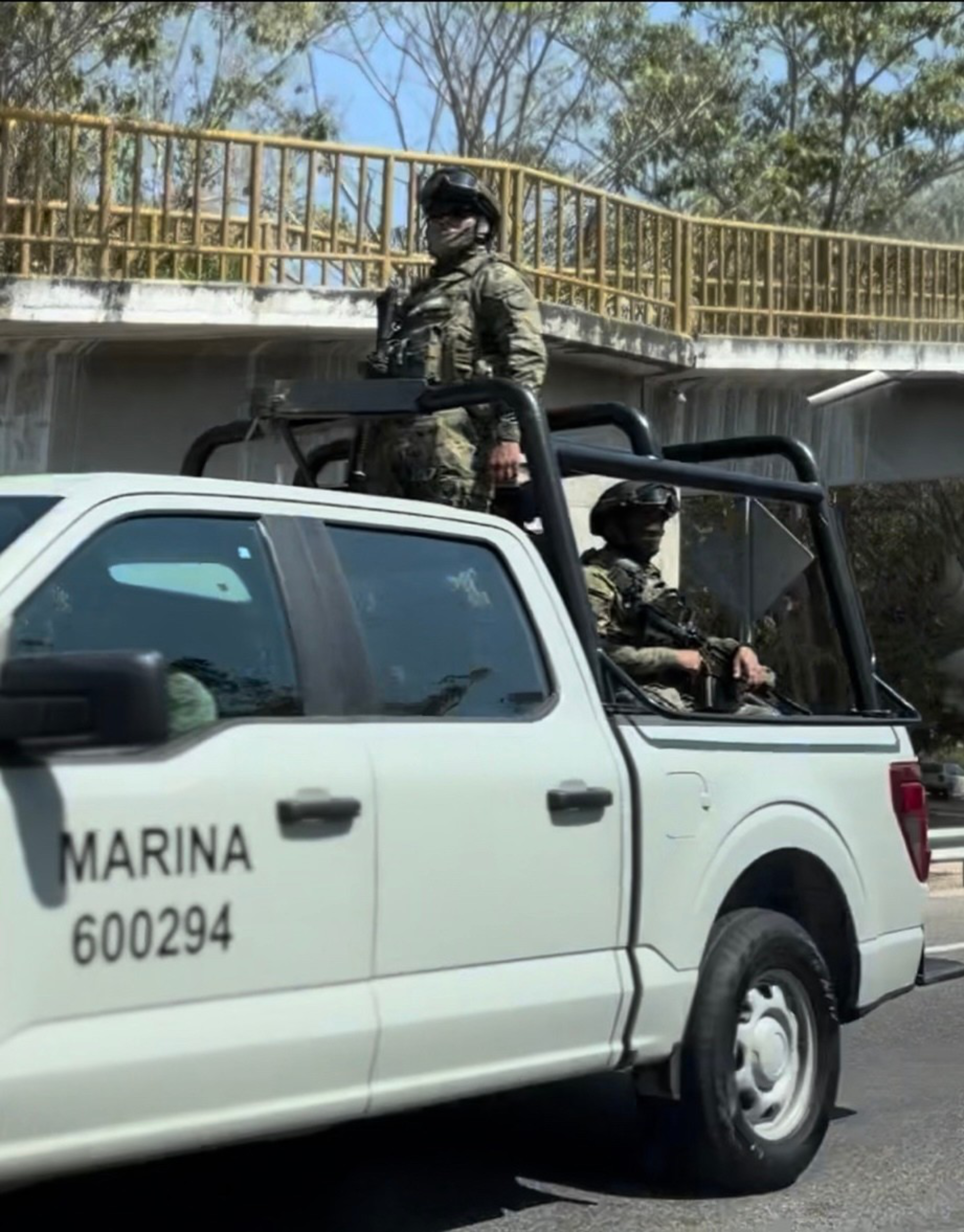 Two armed soldiers in camouflage gear ride in the back of a white pickup truck labeled “MARINA 600294" driving on a road beneath a pedestrian bridge.