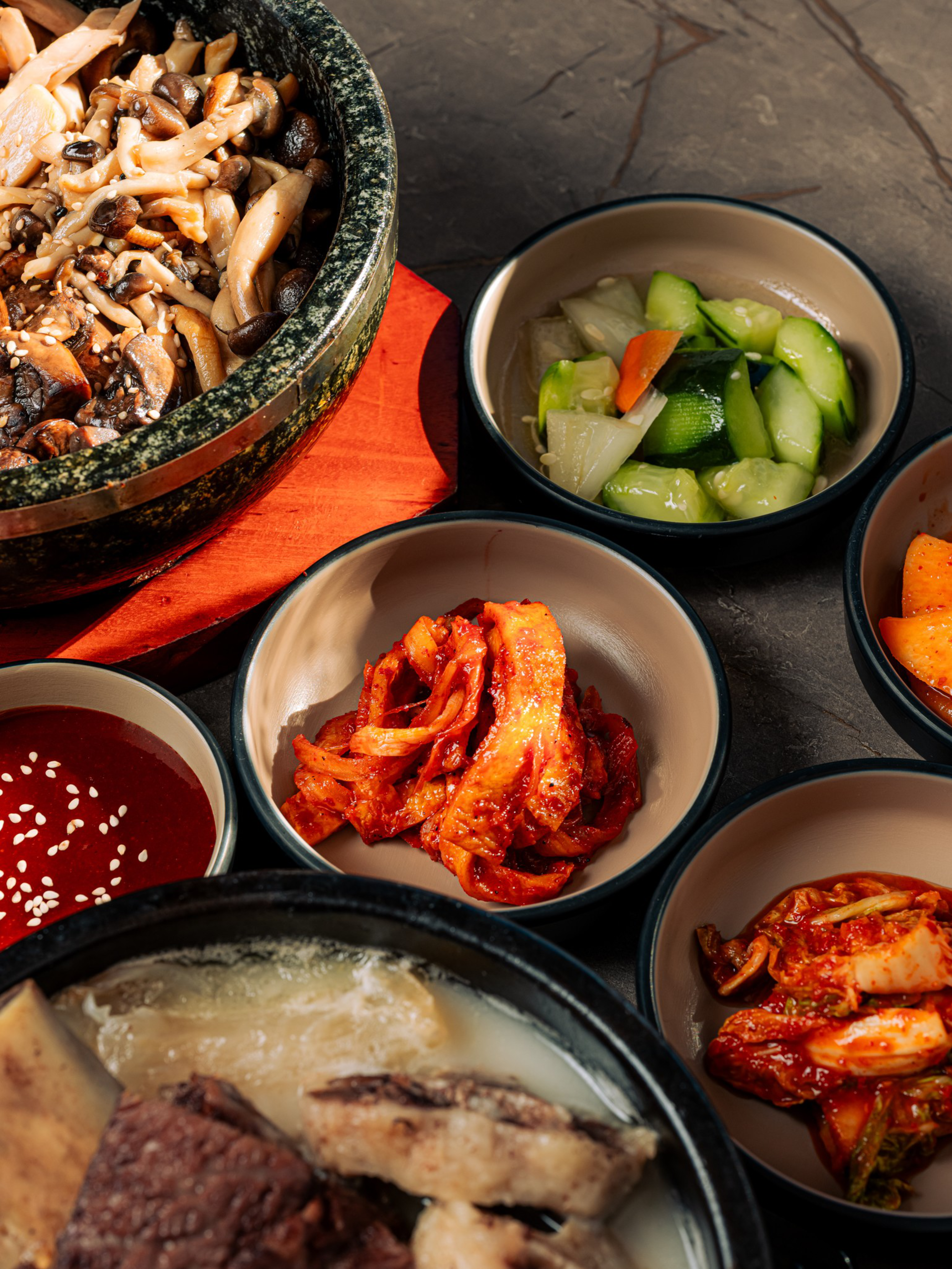 A stone pot with mixed mushrooms, bowls of cucumber salad, two types of kimchi, a red dipping sauce with sesame seeds, and a soup with beef ribs.