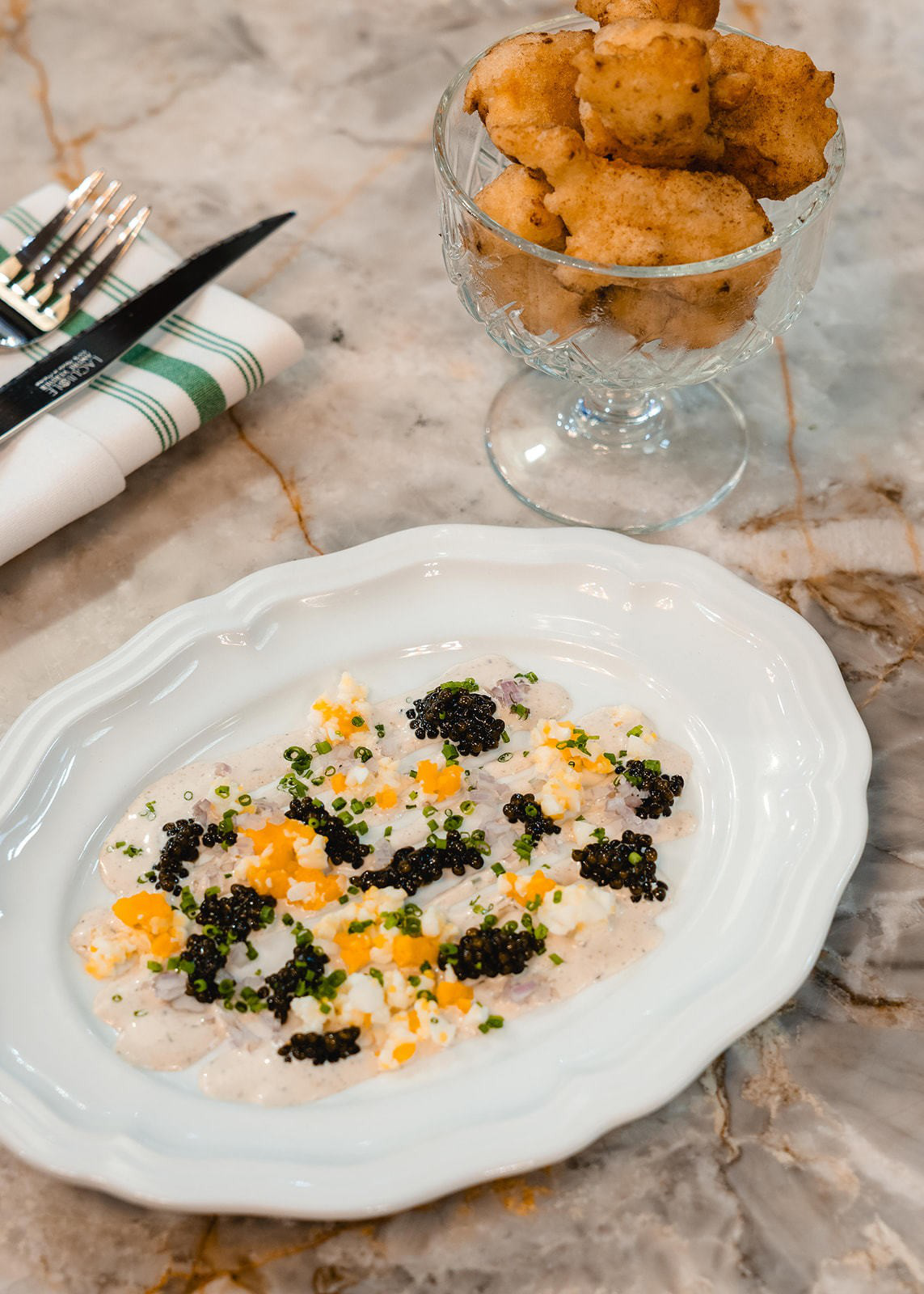 A white plate with a creamy dish topped with black caviar, chives, and orange garnish sits on a marble table next to a glass bowl of fried fritters.