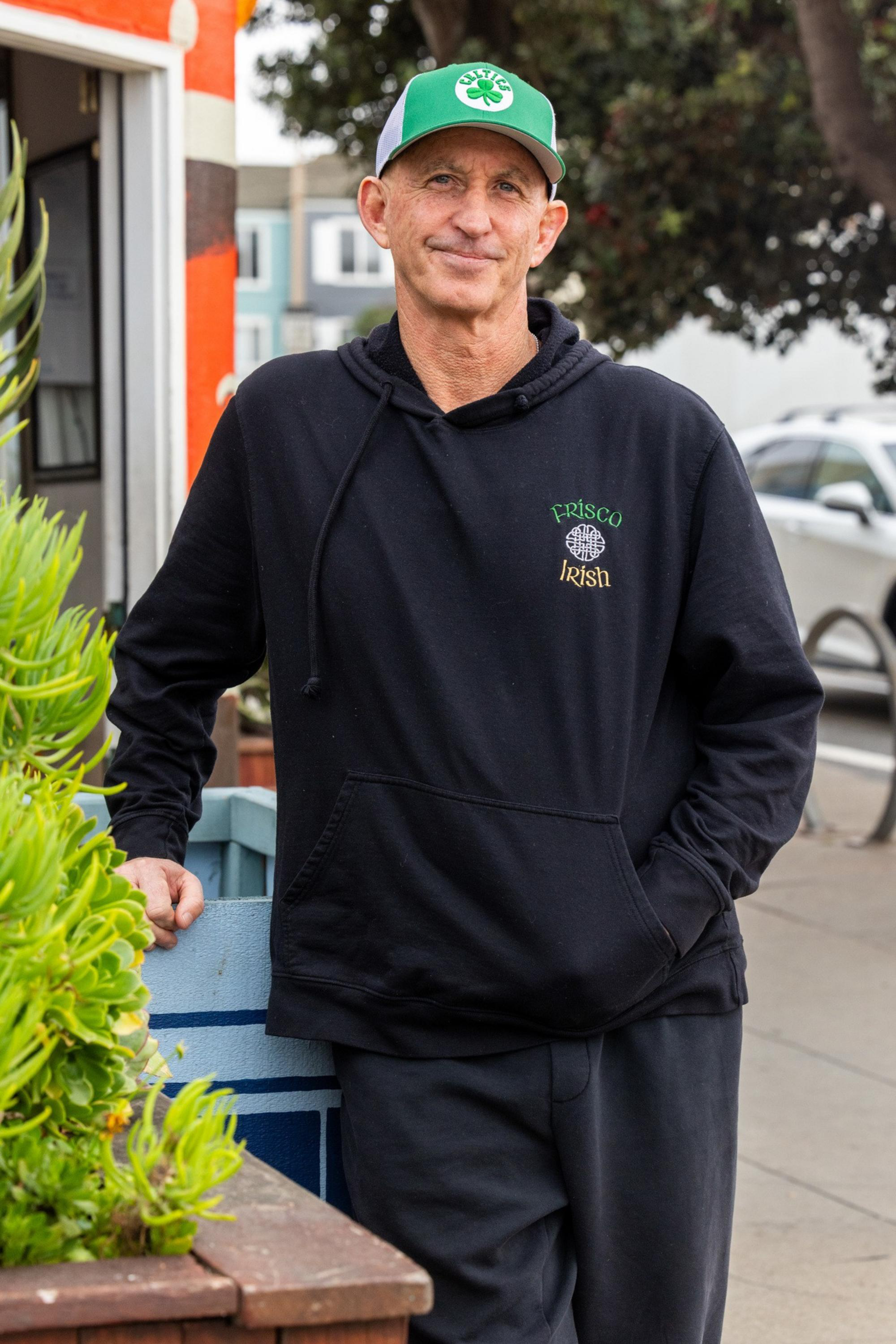 A man in a black hoodie with “Frisco Irish” written on it and a green and white baseball cap leans on a planter box on a sidewalk.