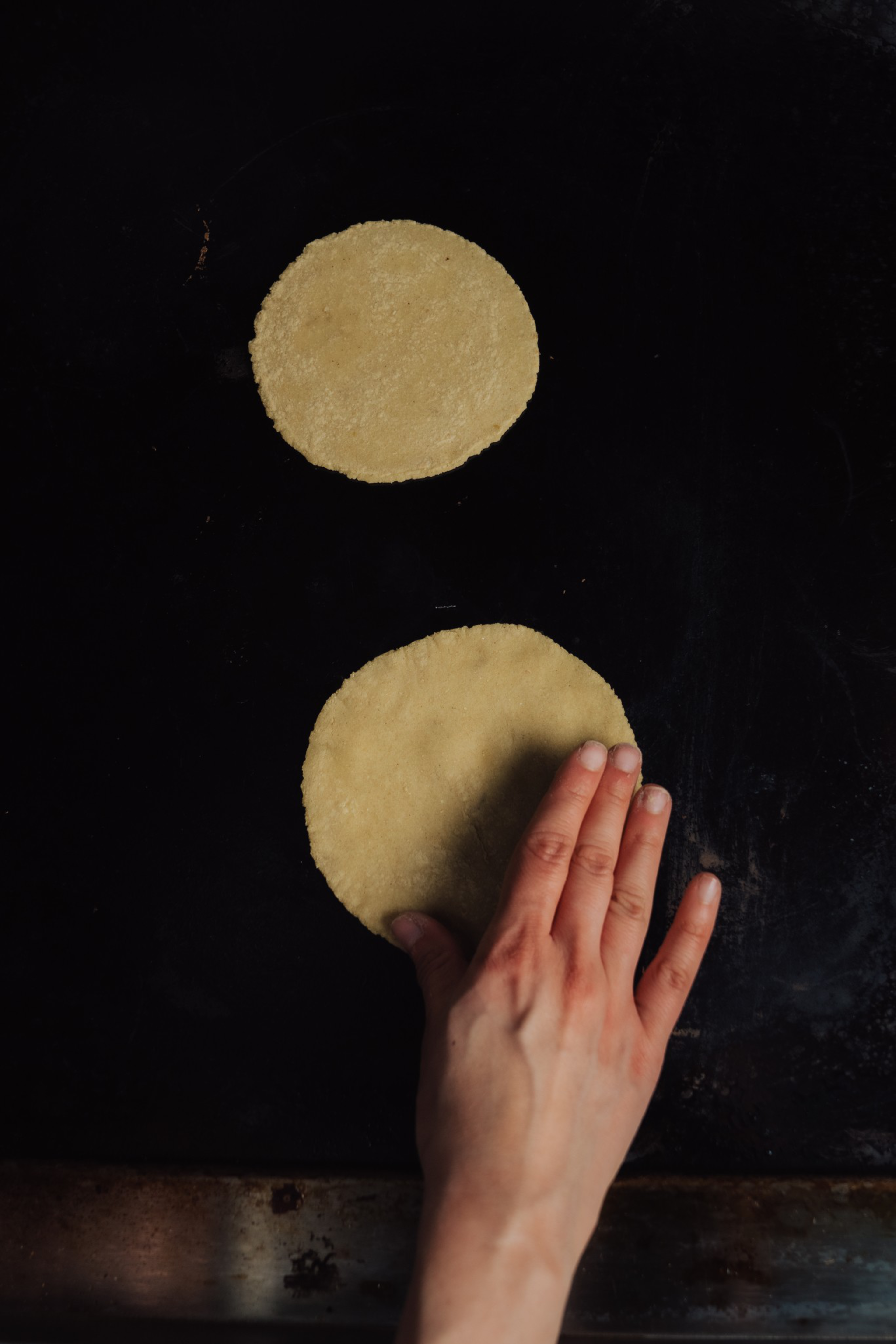 A hand is flattening a round dough disc on a dark surface, with another smaller round dough disc placed above it.