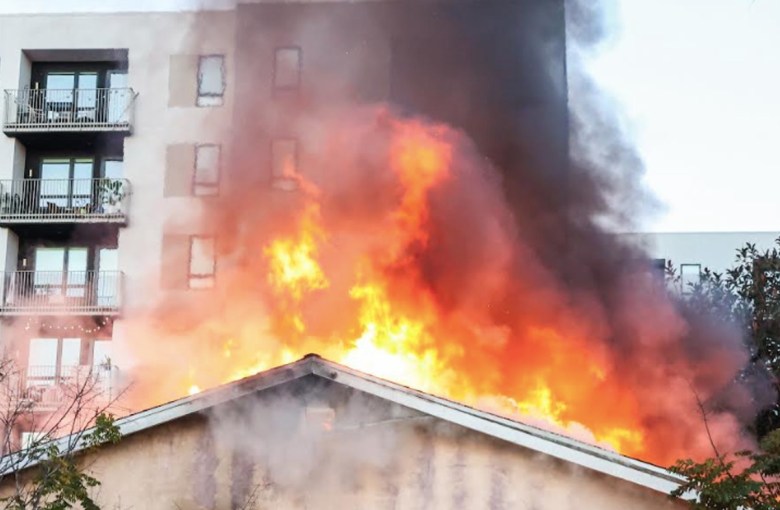 Flames burst from the ceiling of a structure in the shadow of an apartment complex. 