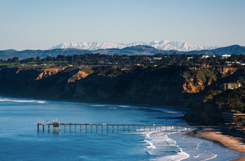 The bright ocean waters on a sunny day around Scripps Pier in La Jolla framed by a view of local terrain and snow in the distance on the mountain tops.