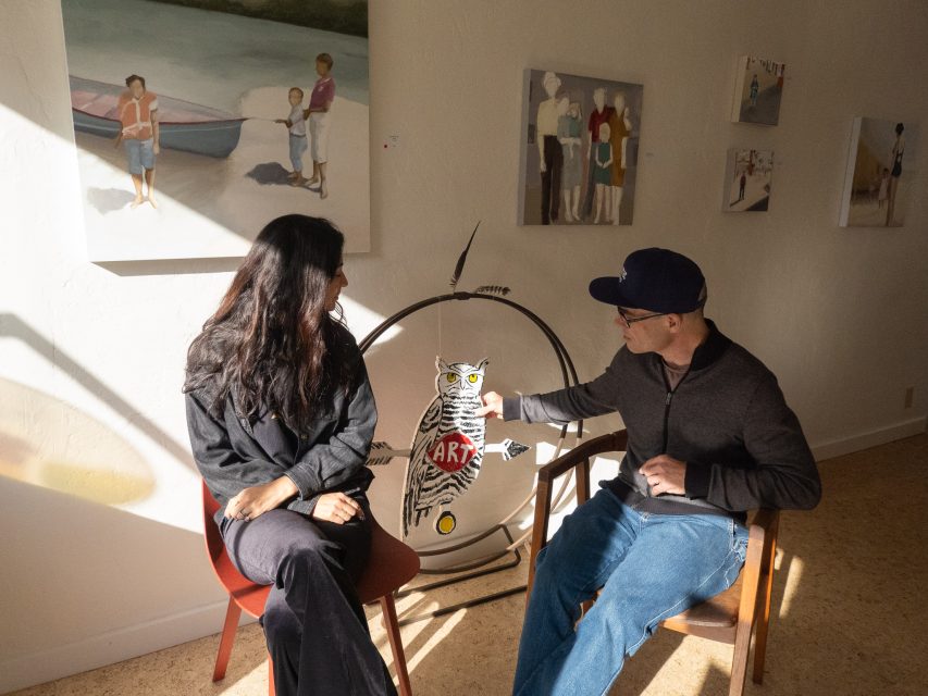 Two people sit on chairs in an art gallery, looking at a painting of an owl with the word "ART" on its chest. Other paintings hang on the walls behind them.