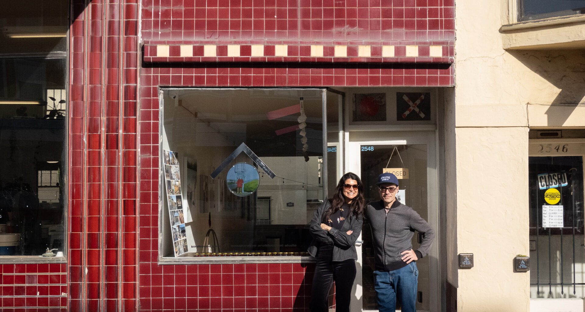Two people stand smiling in front of a storefront with red tiled exterior and a large window, on a sunny day.