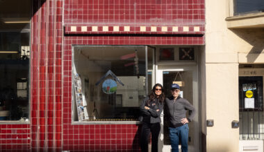Two people stand smiling in front of a storefront with red tiled exterior and a large window, on a sunny day.