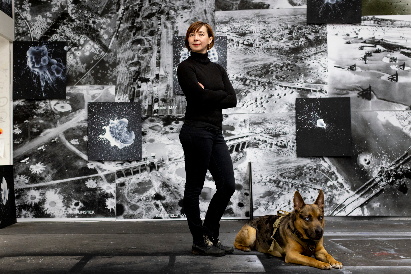 A woman dressed in black fitted top and pants poses with her arms crossed for a full-body portrait with a German shepherd lying at her feet. The woman is standing in front of a black and white collage of aerial images some of which depict explosions.