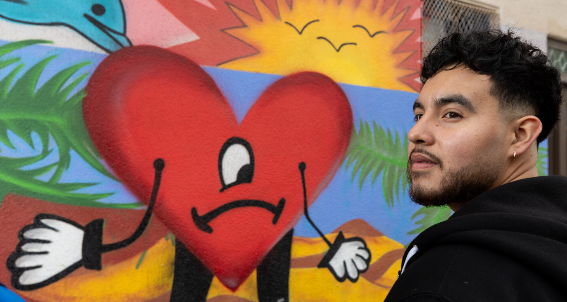 A Latino man with short black hair and a beard, in a black top, stands in front of a colorful mural of a heart with a sad face, a sunset, and dolphins