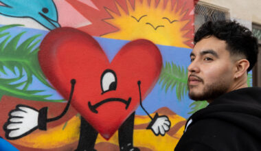 A Latino man with short black hair and a beard, in a black top, stands in front of a colorful mural of a heart with a sad face, a sunset, and dolphins