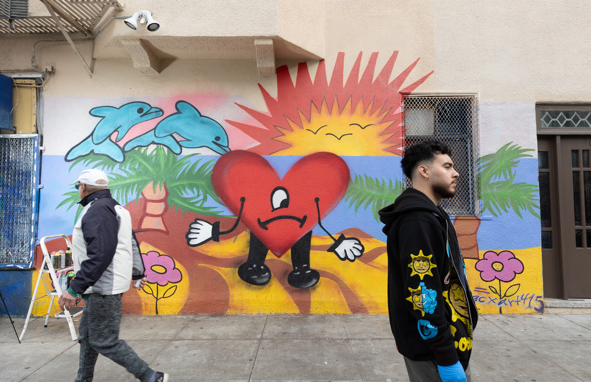 A Latino man with short black hair and a beard, in a black top, stands in front of a colorful mural of a heart with a sad face, a sunset, and dolphins, as an elderly man walks past