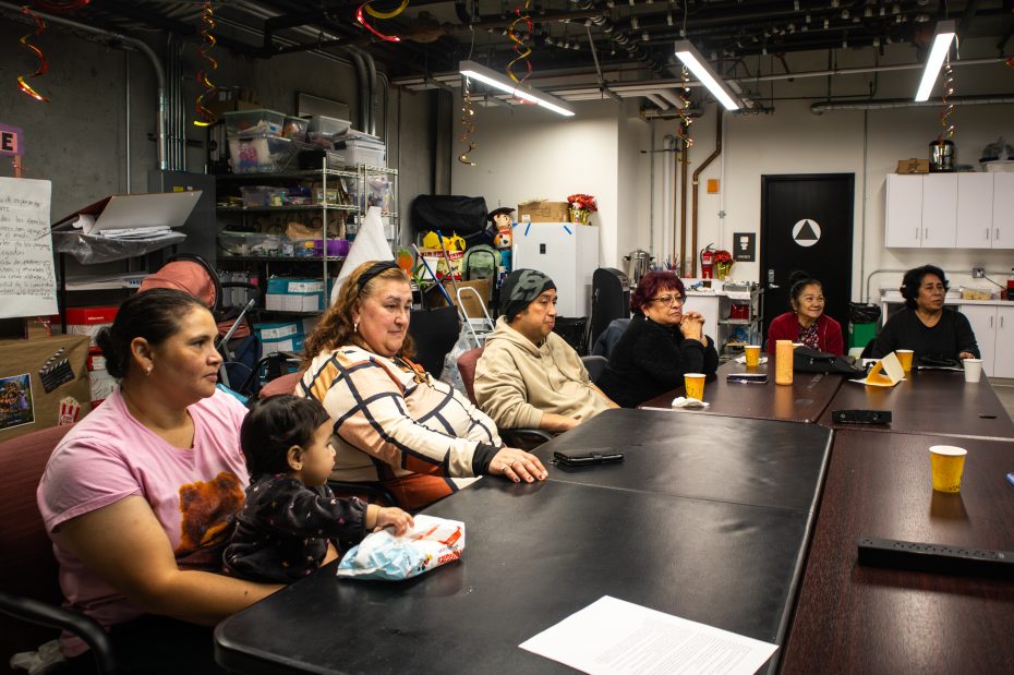 A group of people, including a child, sit around a conference table in a room with supplies and decorations, appearing to listen attentively.