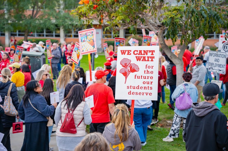 A group of people holding red and white signs stand on a patch of grass outside a school administrative building. One of the main signs in the photo reads, "we can't wait for the future our students deserve."
