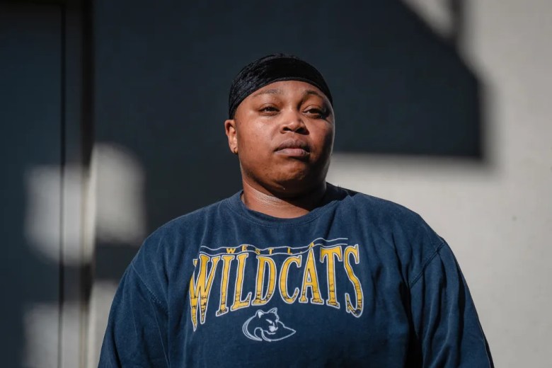A person stands outdoors in bright sunlight wearing a navy sweatshirt with “West Wildcats” printed across the front, looking straight ahead with a serious expression as shadows from nearby structures fall across the wall behind them.