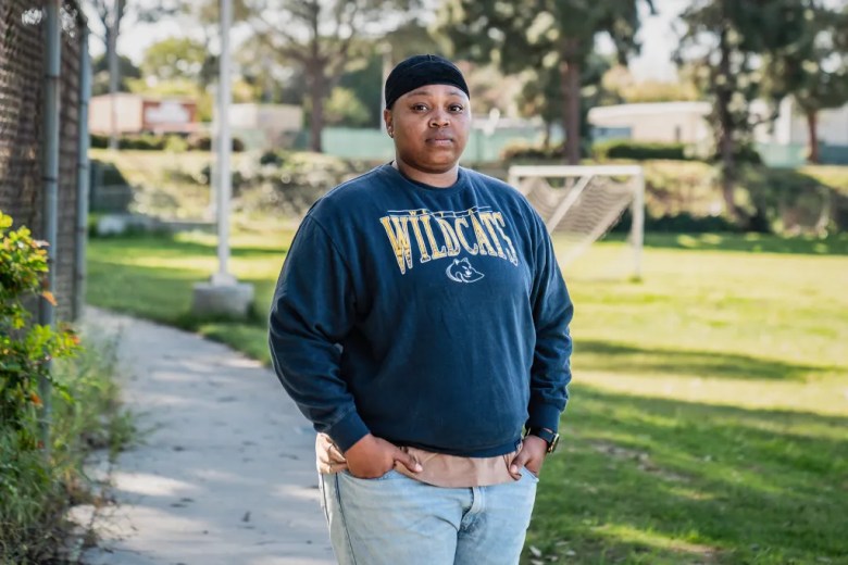 A person stands on a paved path beside a grassy field with a soccer goal in the background, wearing a navy “Wildcats” sweatshirt and light jeans, hands in their pockets, looking calmly toward the camera as afternoon light filters through trees behind them.