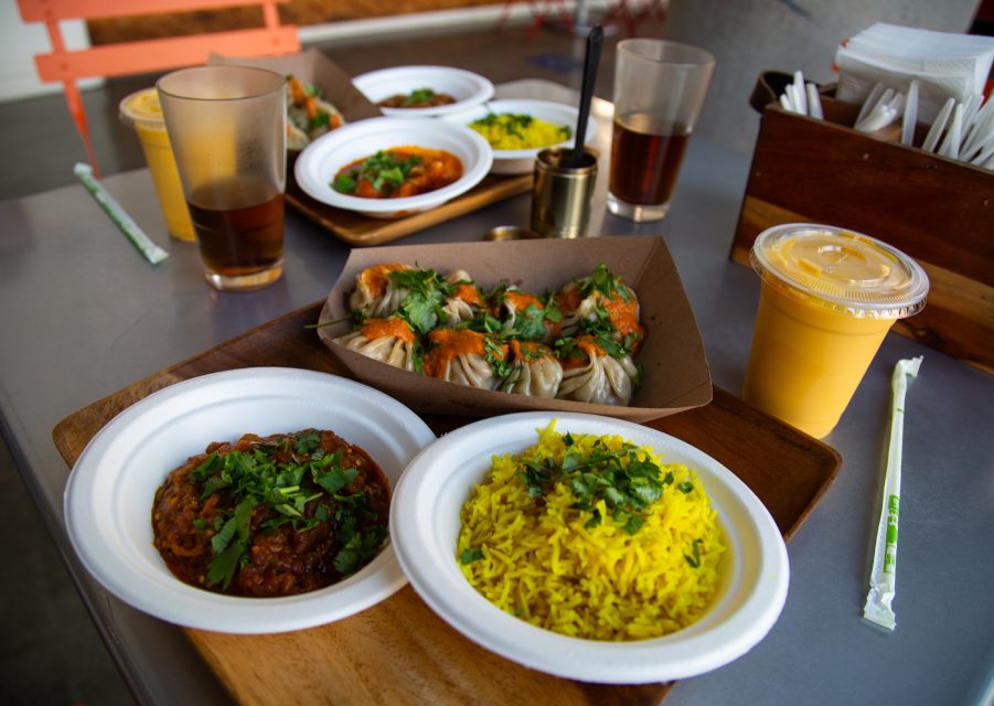 A meal with yellow rice, a curry dish, steamed dumplings with sauce, two glasses of dark drink, and a cup of mango lassi on a table.