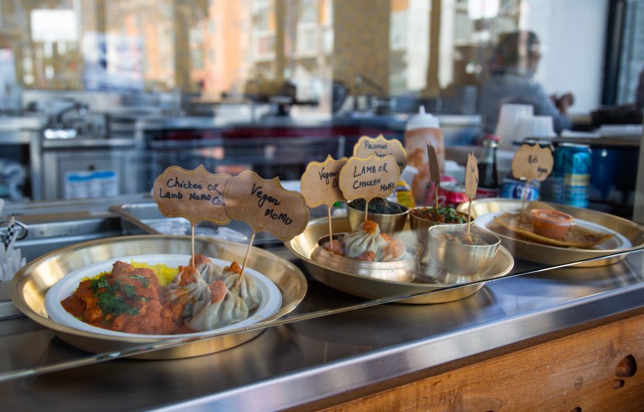 Plates of momos and sides are displayed behind a glass counter, each plate labeled with handwritten signs indicating different flavors like chicken, lamb, and veg.