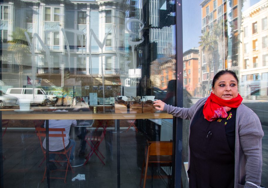 A woman in a red scarf stands outside a glass storefront with a reflection of buildings and a street, pointing to something inside.