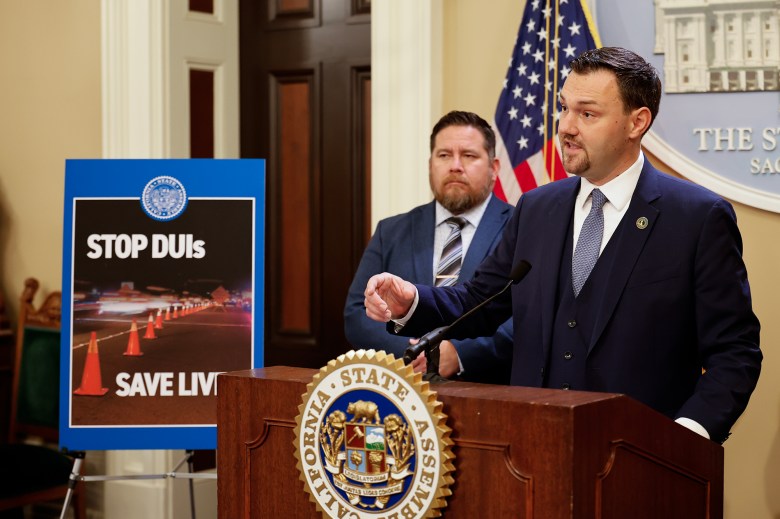 A person in a navy suit speaks at a wooden podium with the California State Assembly seal, gesturing with one hand as another person stands behind them, while a large sign beside the podium reads “STOP DUIs” and “SAVE LIVES” above an image of traffic cones lit by headlights.