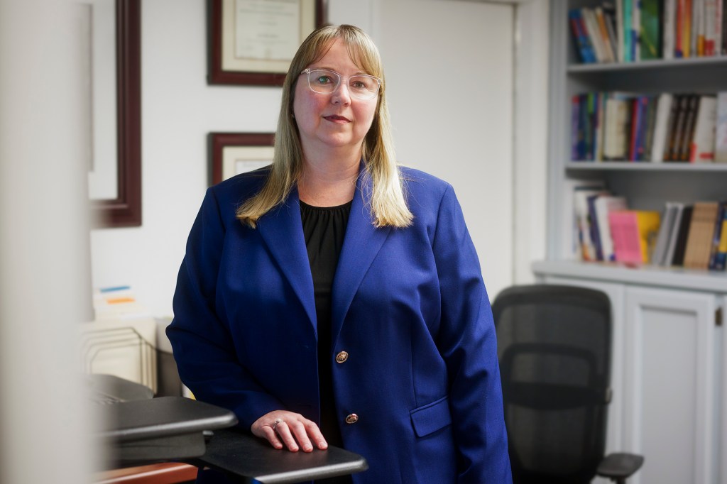 A person wearing glasses and a blue dress jacket stands near a desk inside of an office room with framed degrees in the background on the wall and a bookshelf on the otherside of the frame.