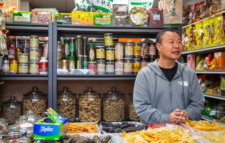 A man stands behind a counter in a shop selling various dried goods and herbs, with shelves of jars and packaged products in the background.