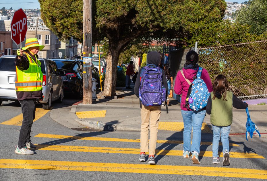 A crossing guard holds a stop sign while three people, including two children with backpacks, cross a street at a crosswalk.