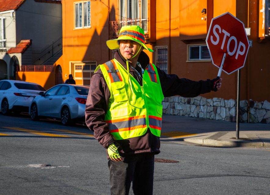 A crossing guard in a neon safety vest and hat holds a stop sign at a street corner with parked cars and orange buildings in the background.