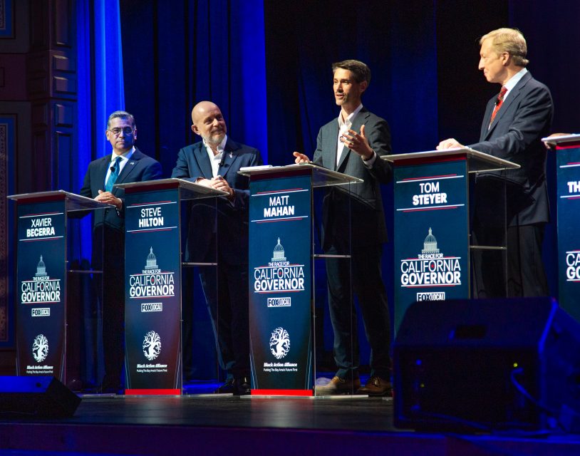 Five men stand at podiums on stage during a California governor debate event, each podium labeled with their names and the event's branding.