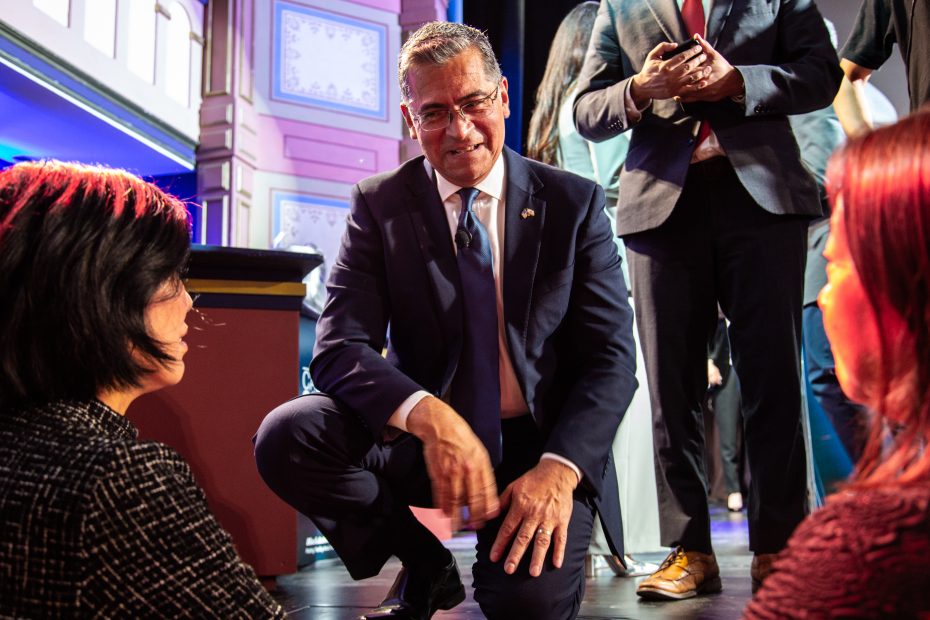 A man in a suit kneels and smiles while talking to two seated women at an indoor event, with other people standing in the background.