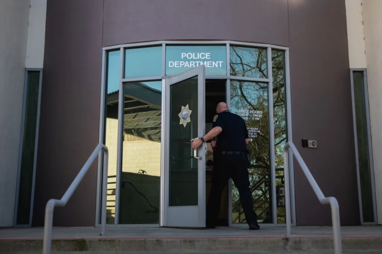 A police officer open the front door of a police deparment's building. The door includes the words "police department" along with the seal of the University of California police.