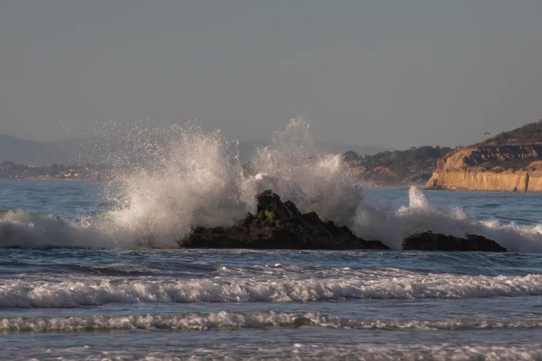 Waves splash against a brown rock with green moss on it.