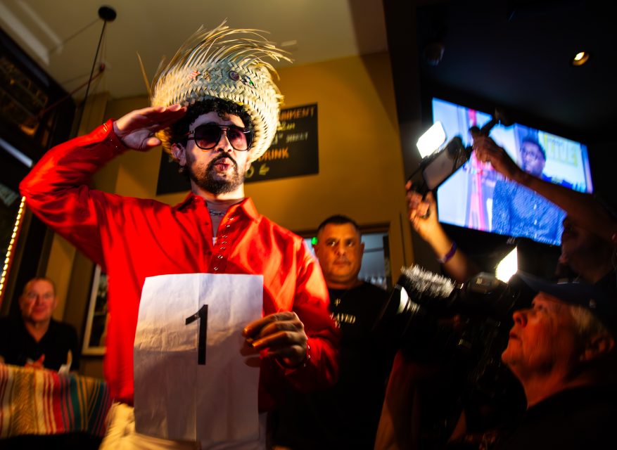 A person in a red shirt, sunglasses, and straw hat salutes while holding a paper with the number 1, surrounded by photographers and onlookers indoors.