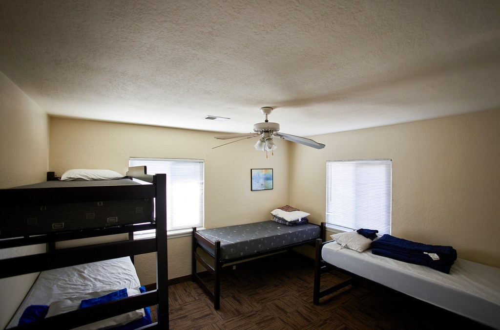 A sparsely furnished bedroom contains three beds: a dark wooden bunk bed at left and two single beds along the walls, each with folded blankets and pillows. Two windows with closed blinds admit soft daylight, and a ceiling fan hangs above. The room has light-colored walls, wood-pattern flooring, and minimal decoration aside from a small framed picture.