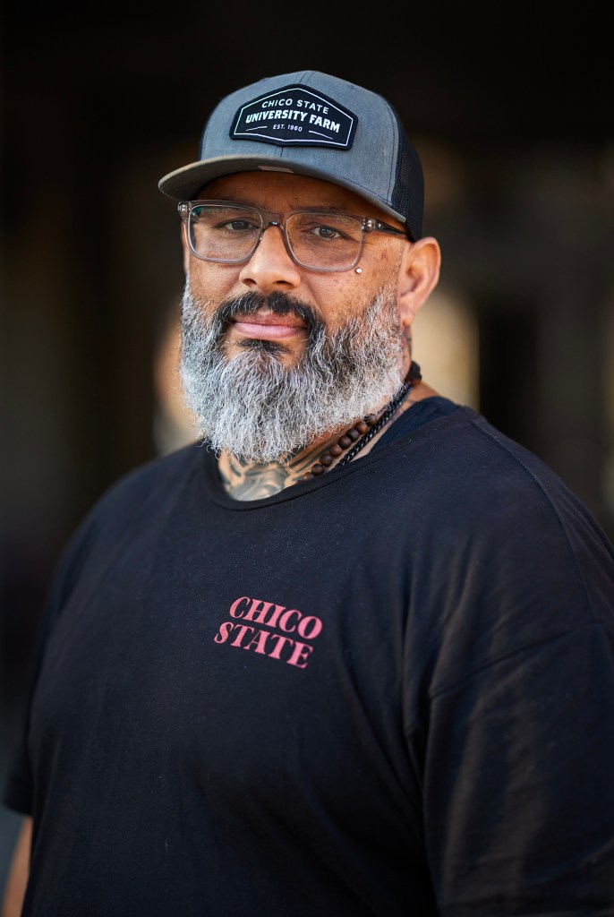 A person with a gray beard and mustache looks toward the camera with a slight smile. They wear glasses, a dark cap that reads “Chico State University Farm,” and a black T-shirt with “Chico State” printed on the chest. A beaded necklace is visible at their collar, and faint tattoos appear on their neck. The background is softly blurred and dark, drawing attention to their face.