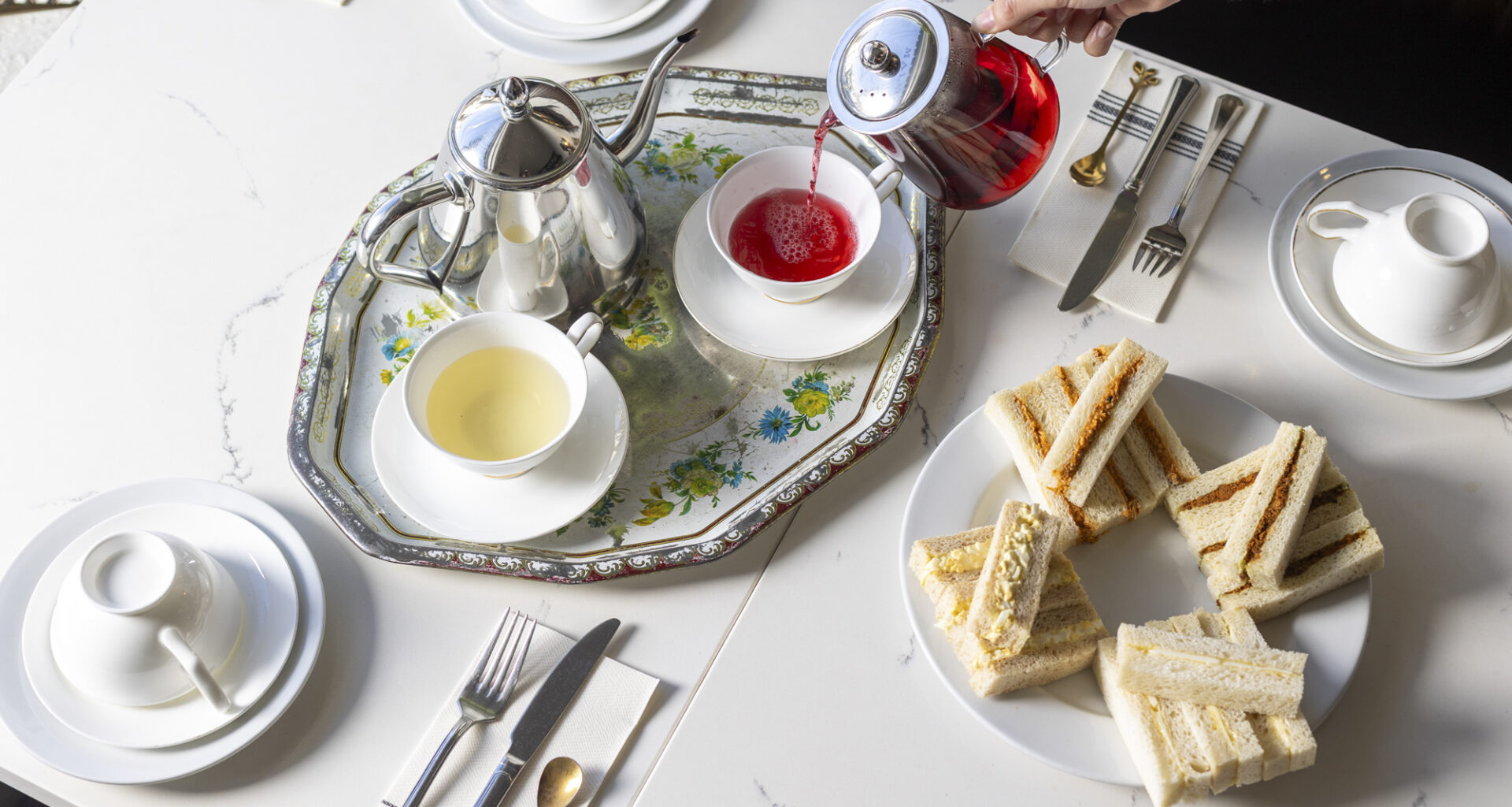 A woman's hand is seen pouring red hibiscus tea into a porcelain tea cup. On the table is a plate of finger sandwiches.