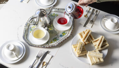 A woman's hand is seen pouring red hibiscus tea into a porcelain tea cup. On the table is a plate of finger sandwiches.