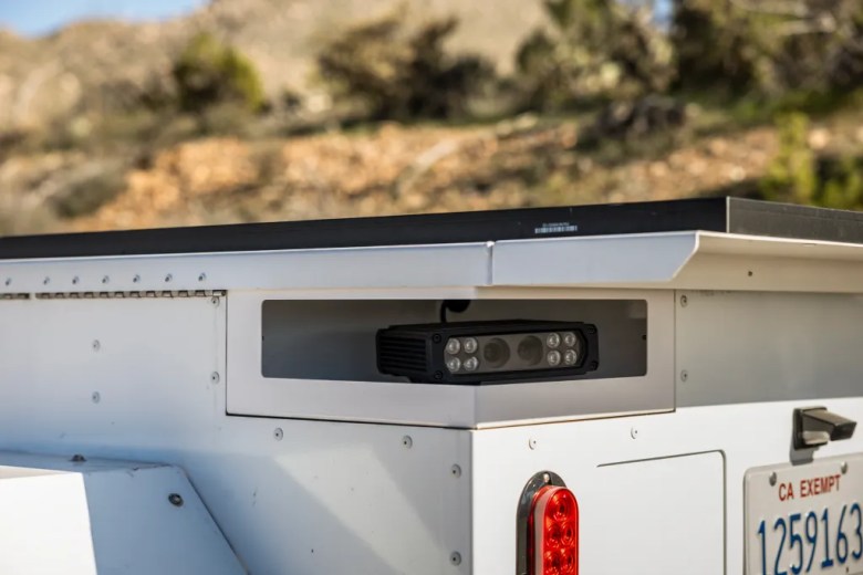Close-up of a camera unit mounted inside a recessed compartment on a white roadside trailer, with rocky hills and vegetation blurred in the background.