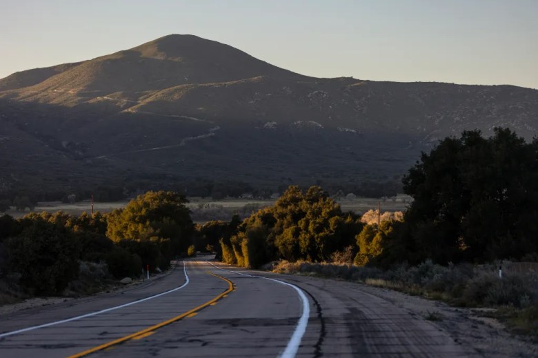 A two-lane road curves through a tree-lined valley toward sunlit mountains, with long evening shadows stretching across the landscape.